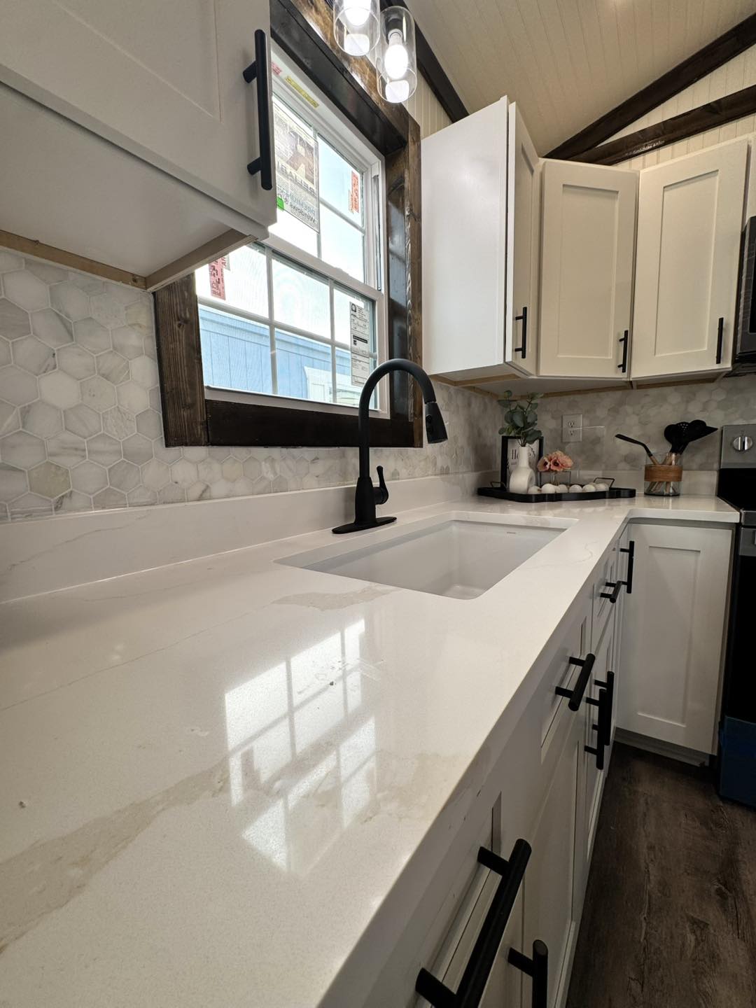 Bright kitchen with white cabinets, black hardware, and a sleek quartz countertop. A matte black faucet complements the light gray hexagonal tile backsplash. Natural light filters through a window, creating a fresh and inviting atmosphere.