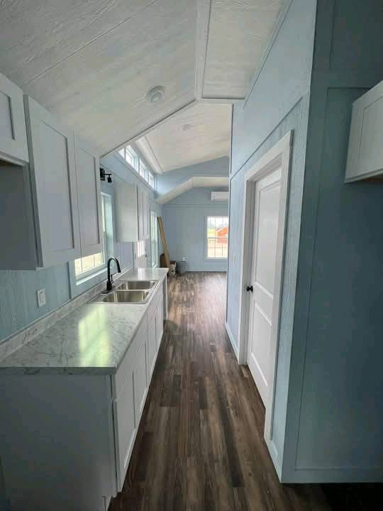 Compact kitchen with light blue walls, sloped ceiling, marble countertop, and dark wood floor. Bright light from large windows creates an airy feel.