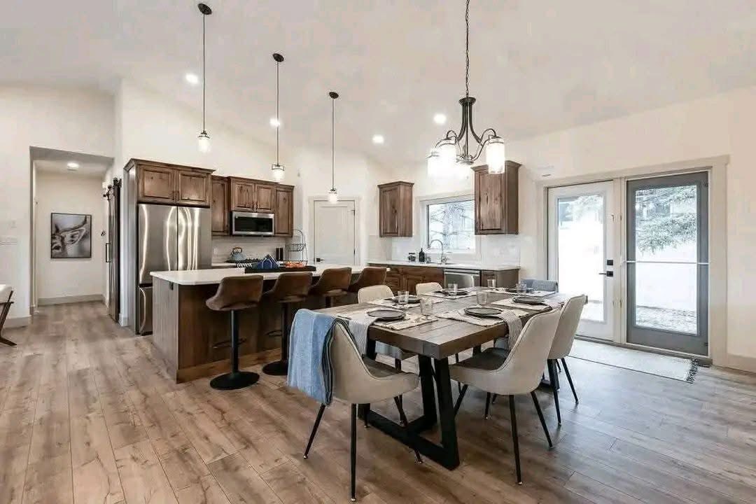 Spacious kitchen and dining area with wooden floors, featuring a large island, pendant lights, brown cabinets, and a set dining table with chairs. Bright and inviting.