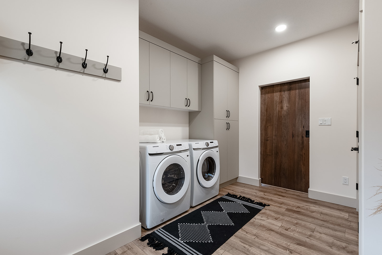 Modern laundry room with white washer and dryer, light gray cabinets, and a wooden door. A black and white rug covers the wood floor, and coat hooks are on the wall.