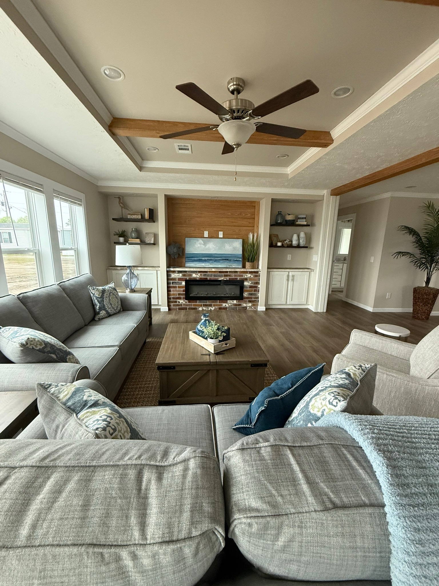 Spacious living room with gray sofas, blue and patterned pillows. Wall-mounted TV above a brick fireplace. Wooden beams and ceiling fan add warmth. Cozy ambiance.