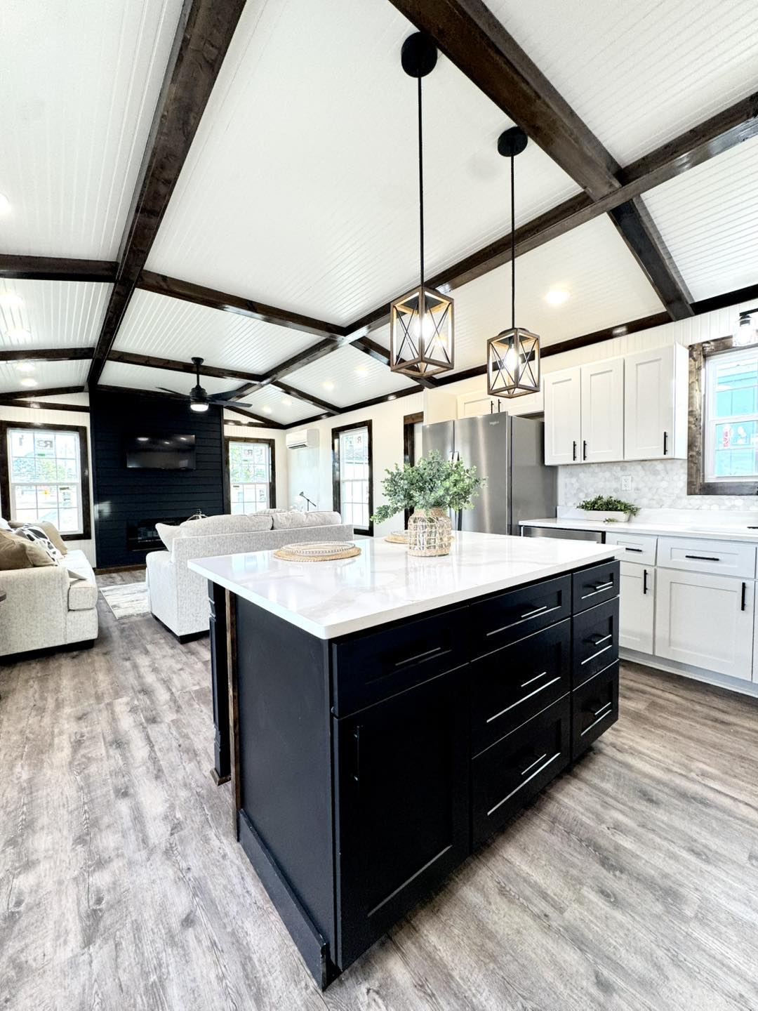 A modern kitchen with high ceilings and exposed beams. Features a large island with dark cabinets and white countertop, pendant lights, and bright natural light.