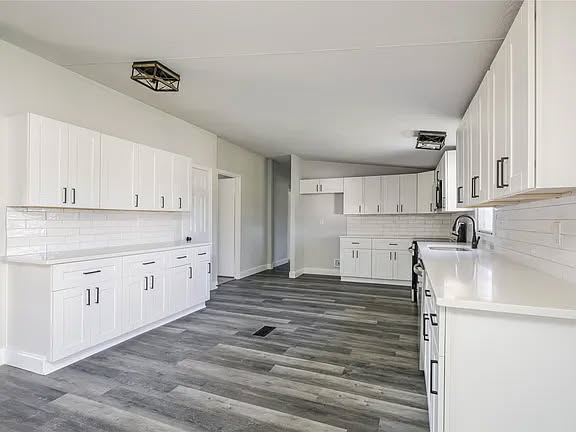 Spacious, modern kitchen with white cabinetry, dark handles, and gray wood flooring. Bright and clean ambiance with geometric light fixtures.