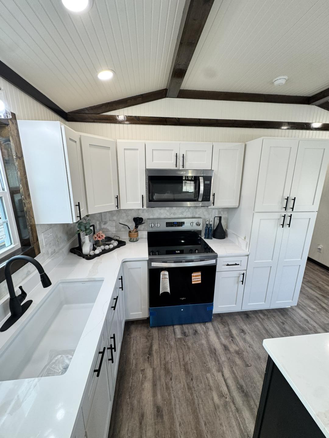 Modern kitchen with white cabinets, black handles, and stainless steel appliances, including a microwave and oven. Wooden flooring and ceiling add warmth.