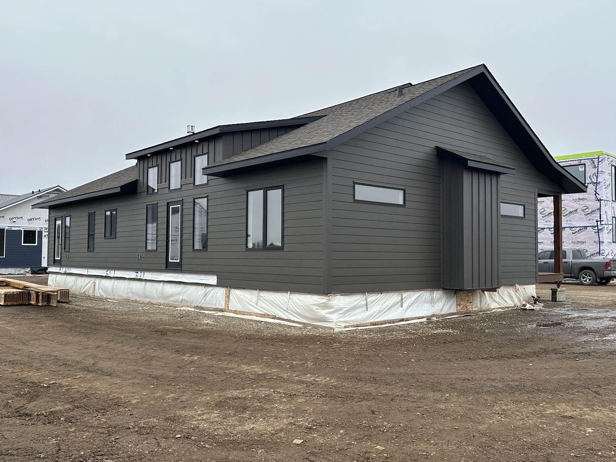 Single-story, modern, dark gray house under construction, set on a muddy lot. The building features large windows, a sloped roof, and a partially wrapped foundation. Overcast sky suggests a calm but dreary day.