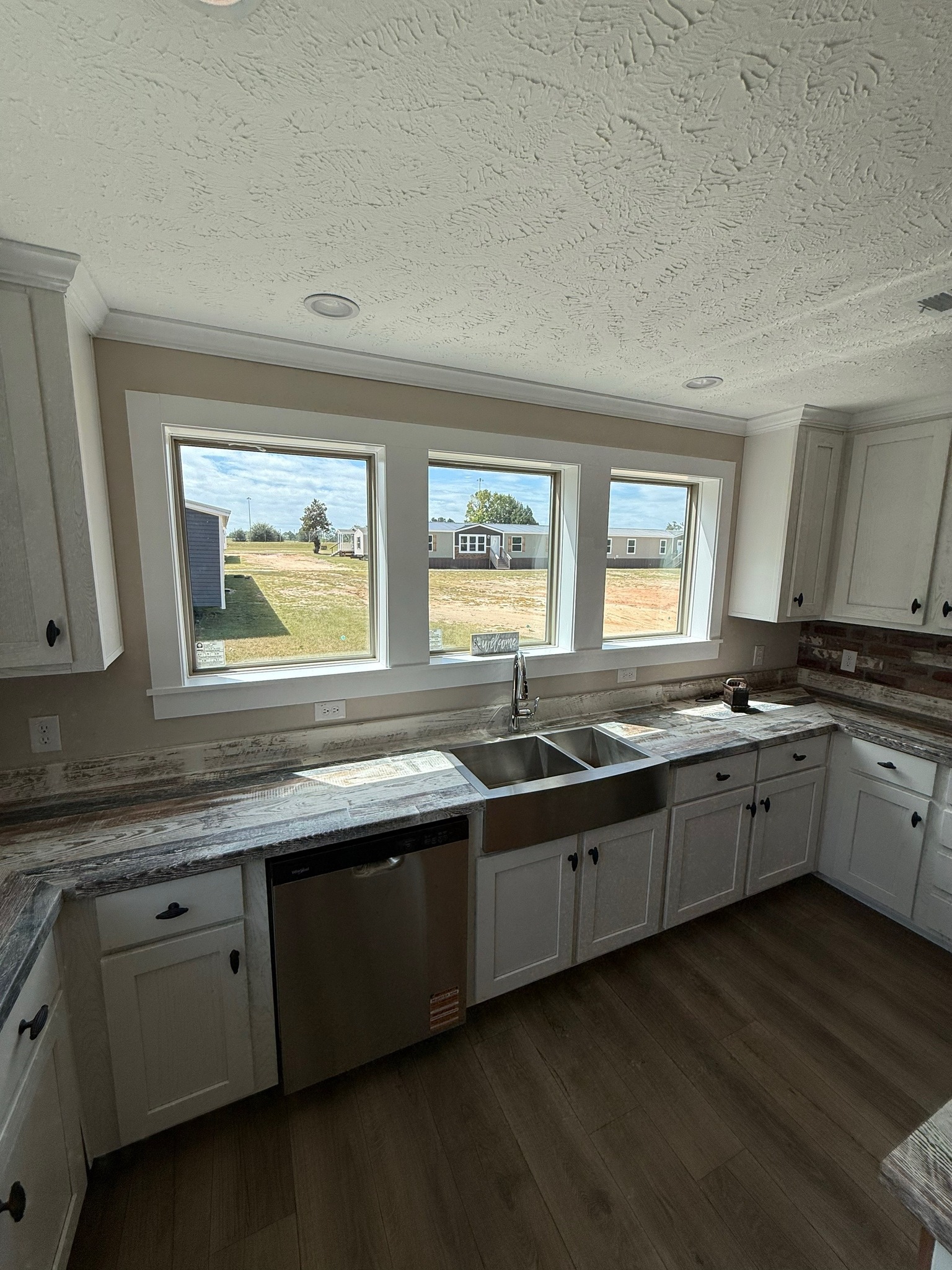 Bright kitchen with three tall windows overlooking a sunny yard. Features white cabinets, stainless steel appliances, and a patterned countertop. Cozy and inviting space.