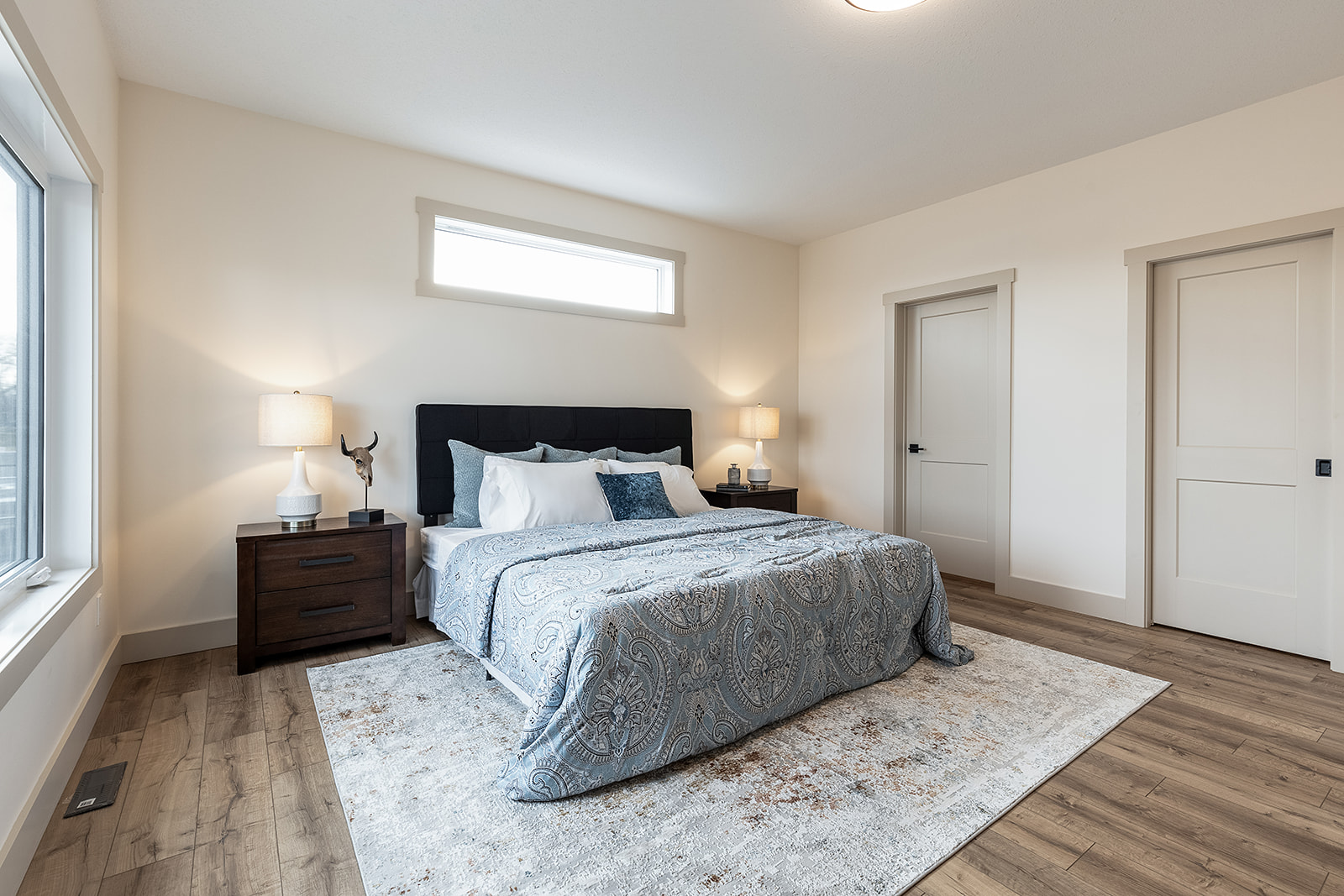 Stylish bedroom with a cozy bed featuring a blue patterned comforter, two dark wood nightstands with lamps, and a soft rug on wooden flooring. Bright and serene ambiance.