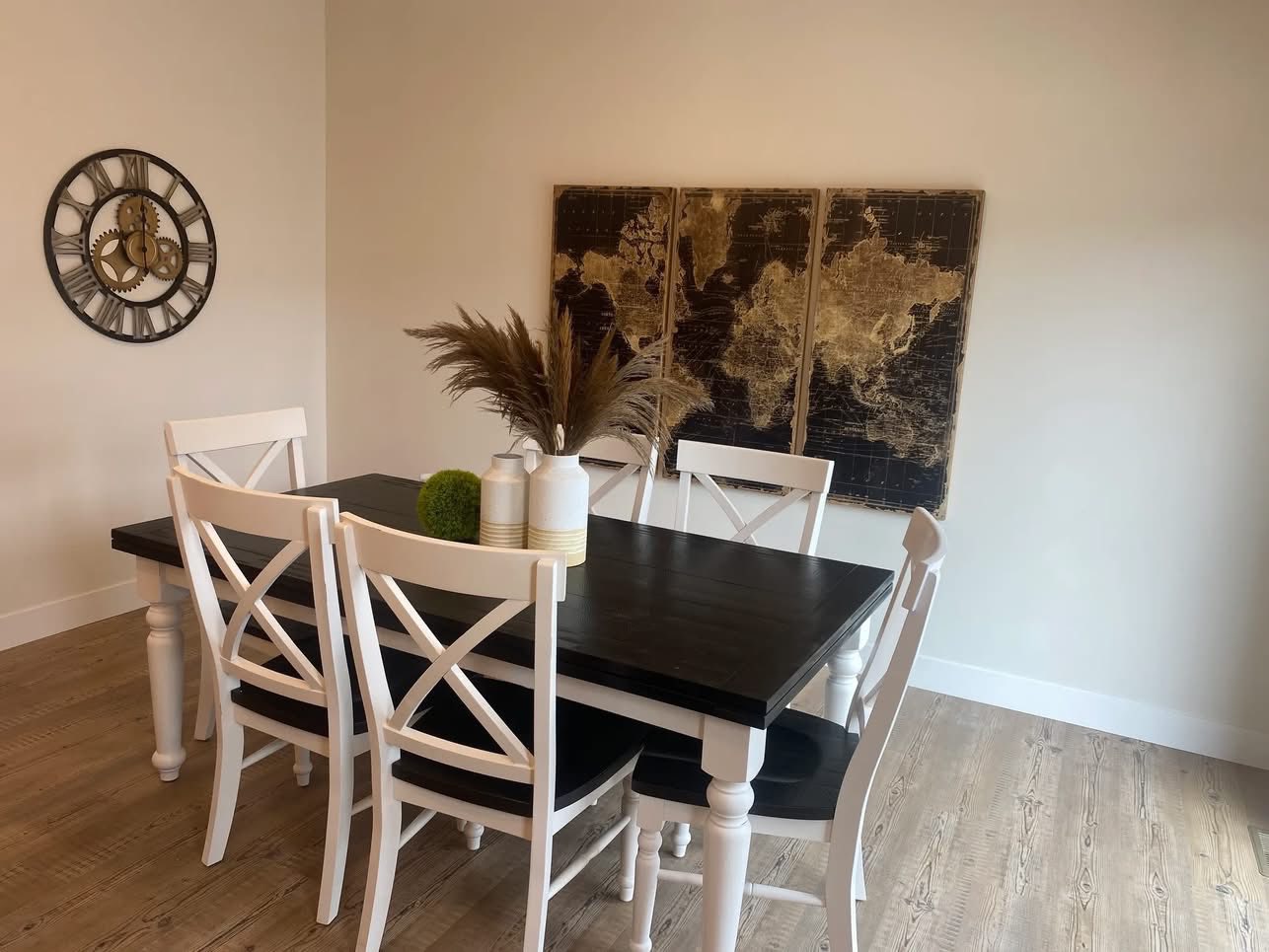 A modern dining room with a black table and four white chairs on wooden flooring. A large world map art piece and a clock adorn the beige walls.