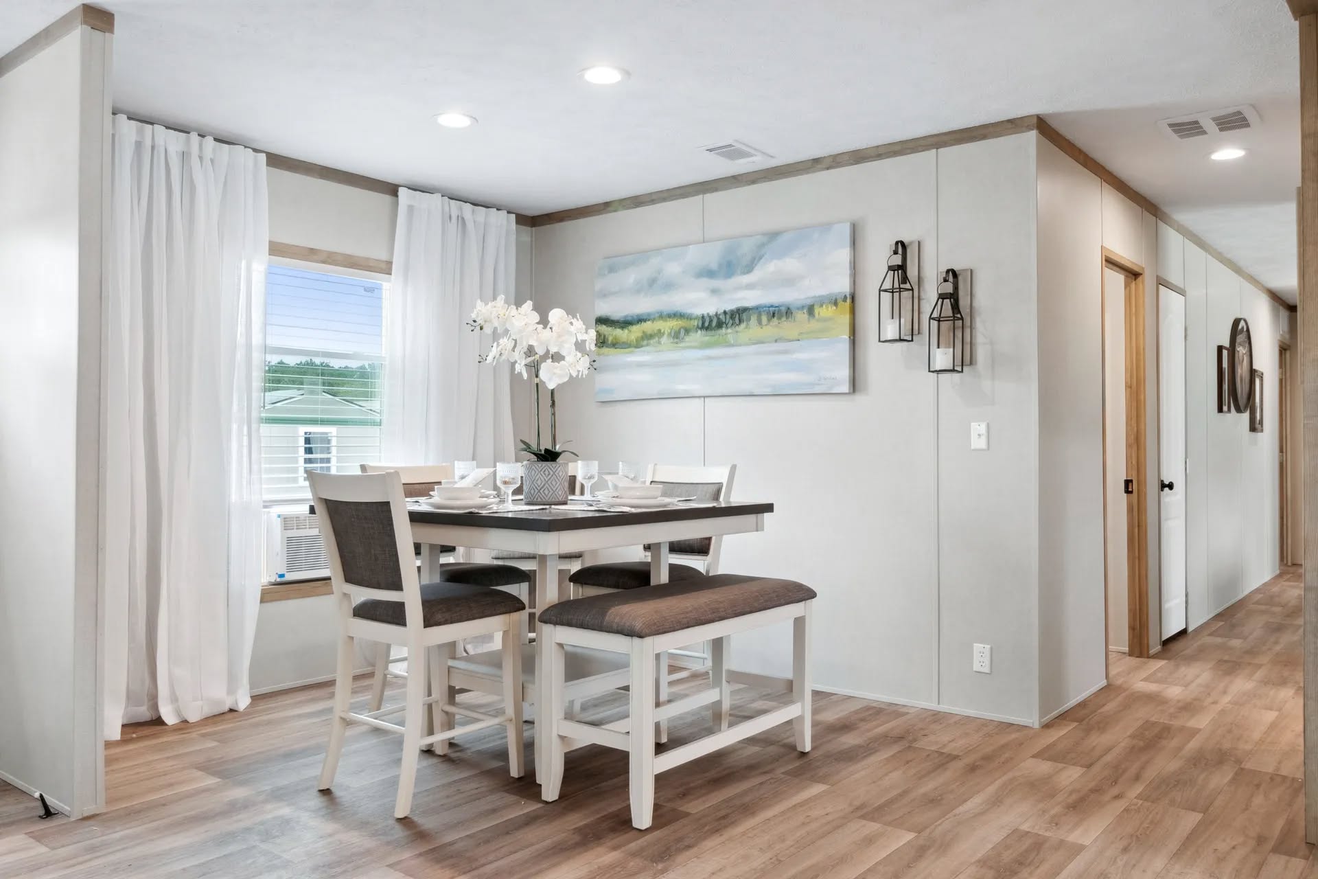 A modern dining room with a wooden table set for four, featuring white and gray chairs and a bench. An orchid centerpiece, abstract landscape painting, and soft lighting create a calm ambiance.
