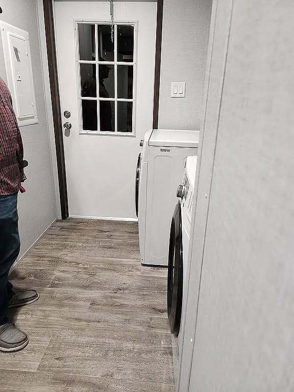 A narrow laundry room with a white door at the end, featuring a window. Two white laundry machines are on the right. A person stands on the left.