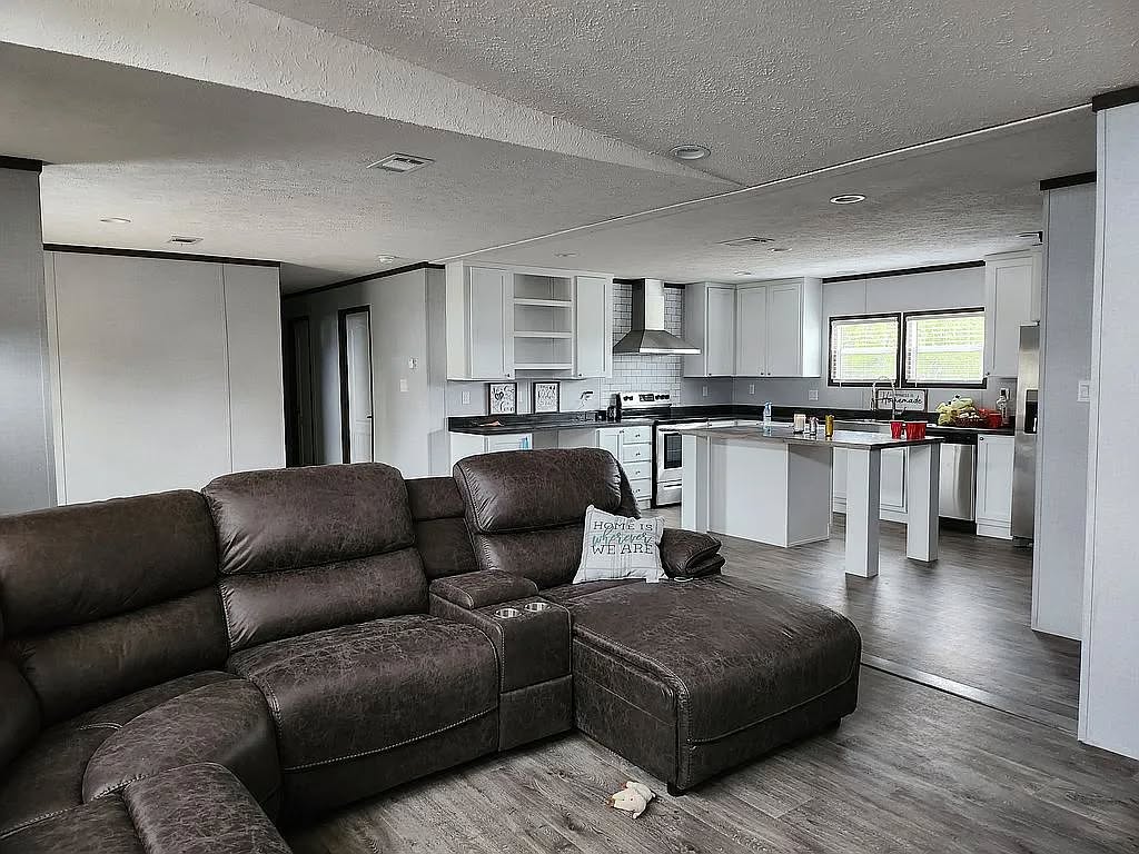 Cozy living room with a dark brown sectional sofa facing a modern white kitchen. Soft light gives a warm, inviting feel. A pillow reads "Home Sweet Home."