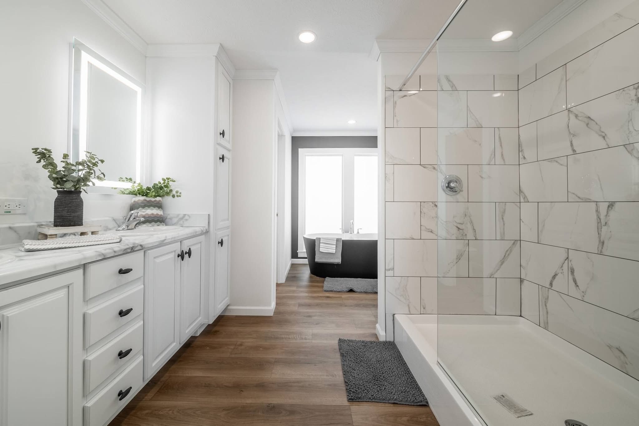 Modern bathroom with wood floors, white cabinets, a glass-enclosed shower with marble tiles, and a black bathtub in the background. Bright and airy ambiance.