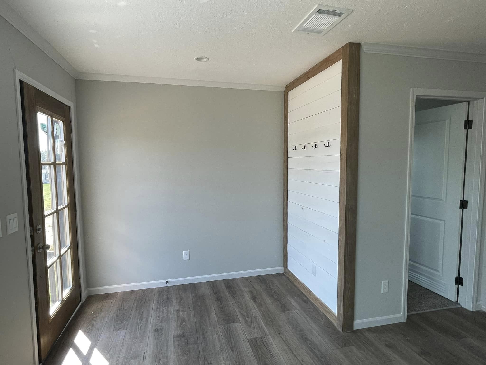 A bright room with light gray walls, wooden flooring, a row of hooks on a white paneled wall, and a glass door on the left. The atmosphere is clean and modern.