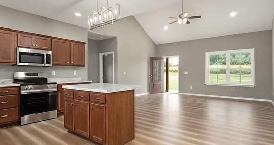 Spacious open-concept kitchen and living area with wooden floors. Modern kitchen features dark wood cabinets, stainless steel appliances, and island. Natural light streams through large window and glass door, creating a welcoming atmosphere.