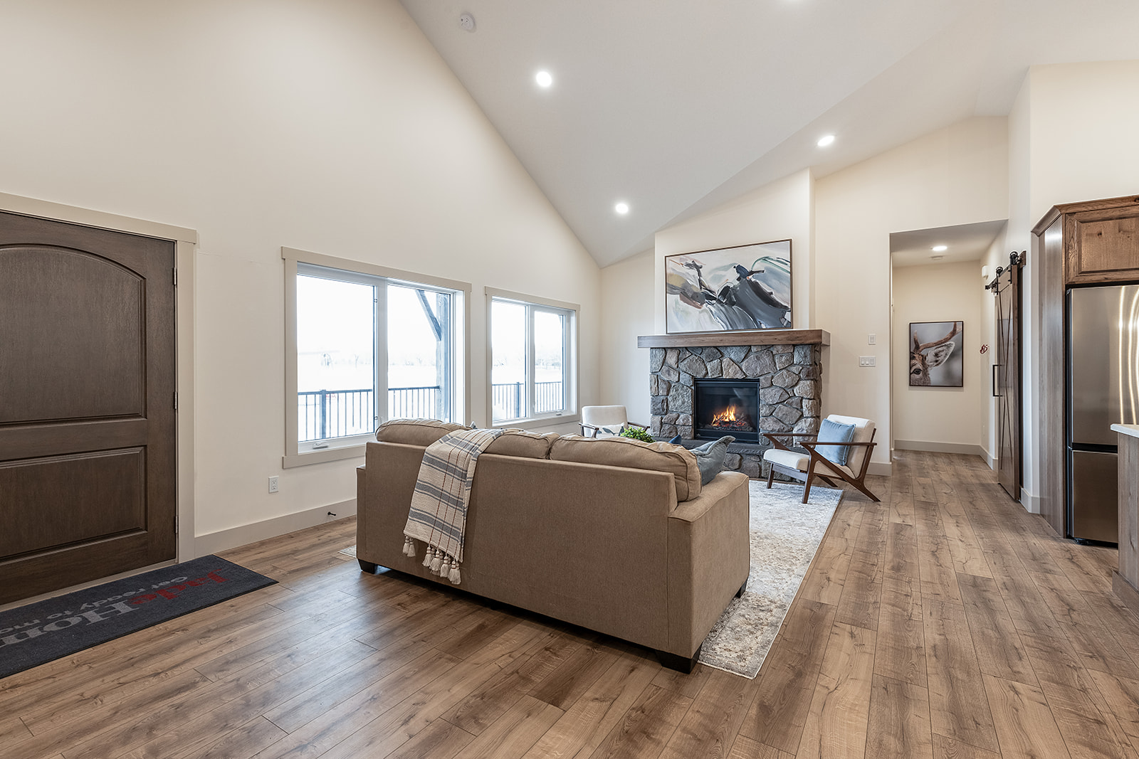 Modern living room with vaulted ceiling and warm tones. Features a stone fireplace, beige sofa with a plaid throw, wooden floors, and bright natural light. Cozy, inviting atmosphere.