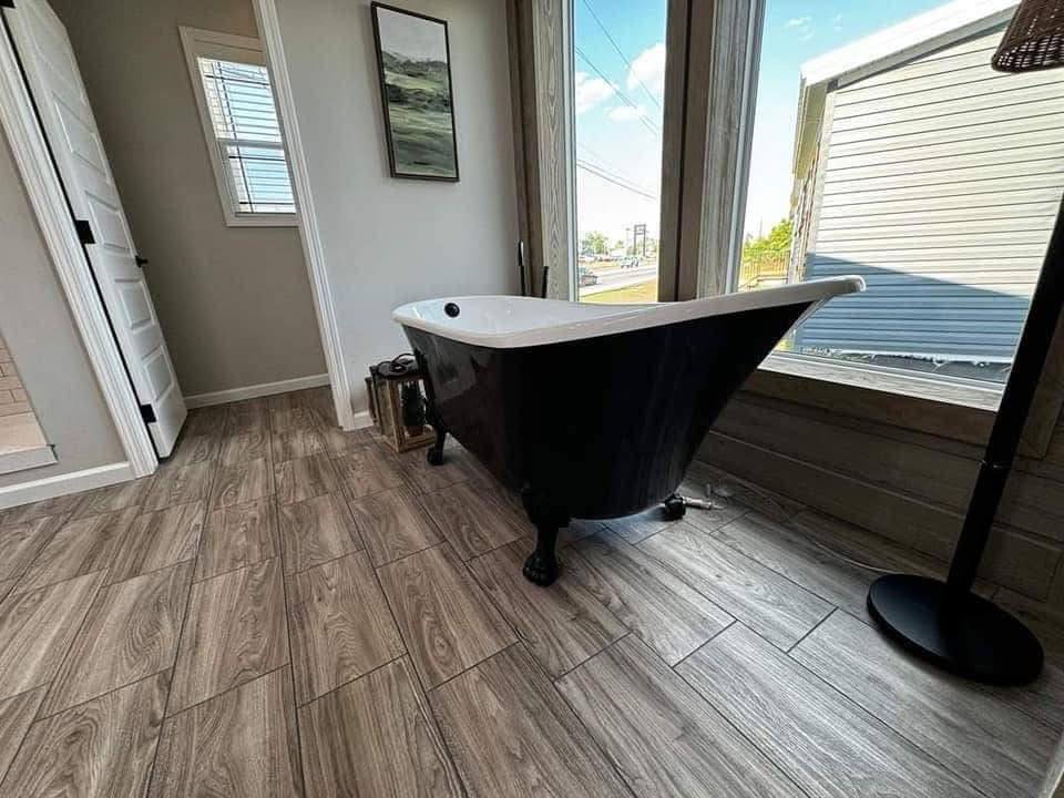 Elegant bathroom with a black clawfoot tub near large windows. Light floods in, illuminating wood-style tiles and creating a tranquil, cozy atmosphere.