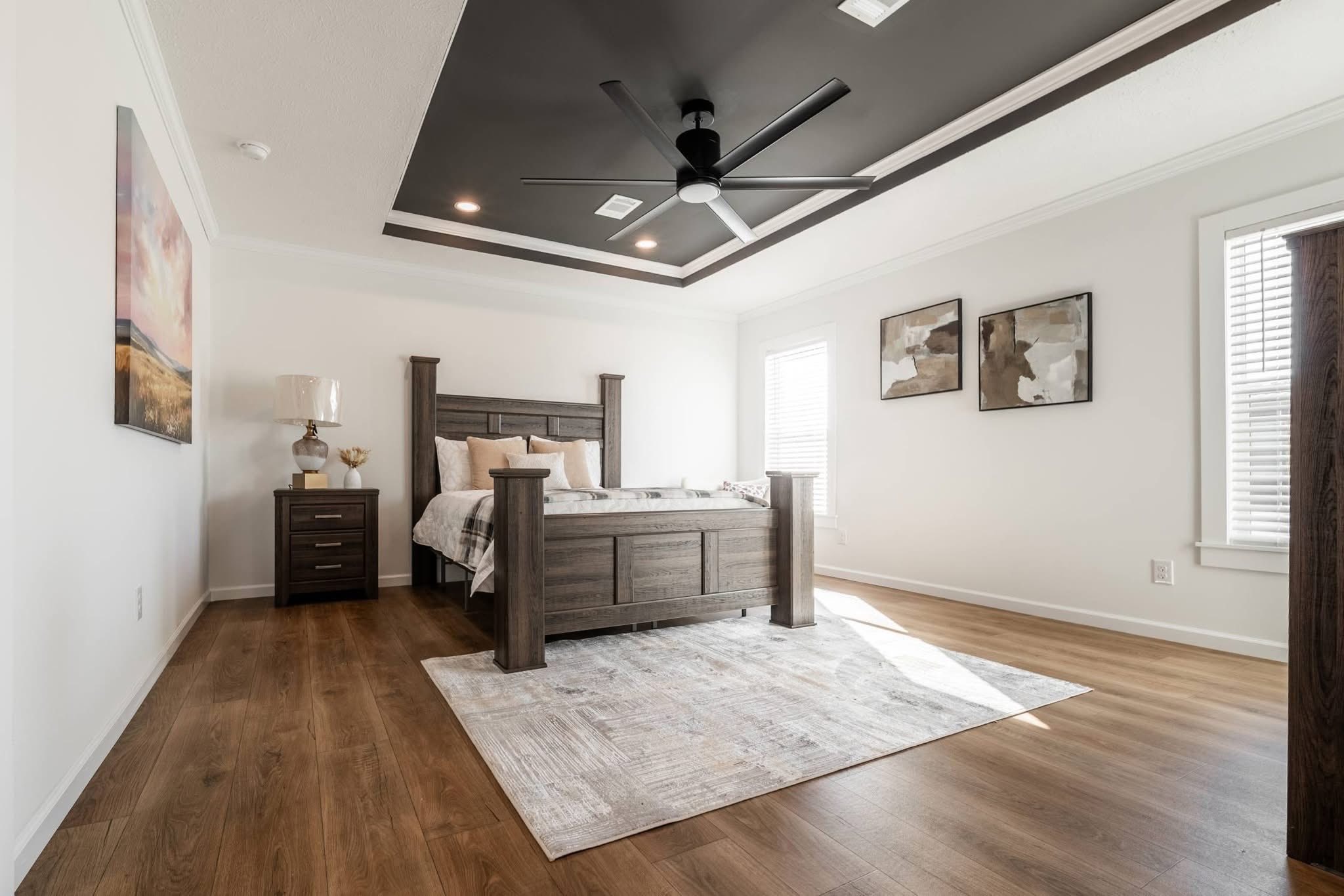 Modern bedroom with a dark wood bed, beige bedding, and a textured rug. Black ceiling with fan, abstract art, and soft light create a serene feel.