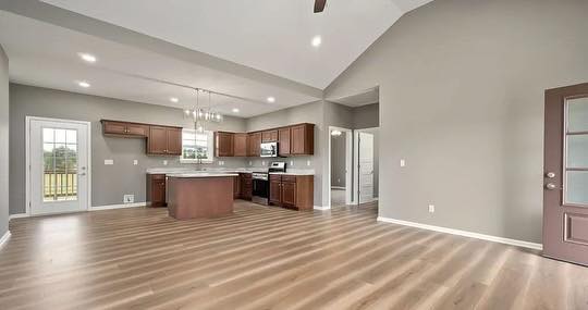 Modern open-plan kitchen and living area with light wood flooring, gray walls, and high ceilings. Wooden cabinets, central island, and large windows provide a bright, airy feel.