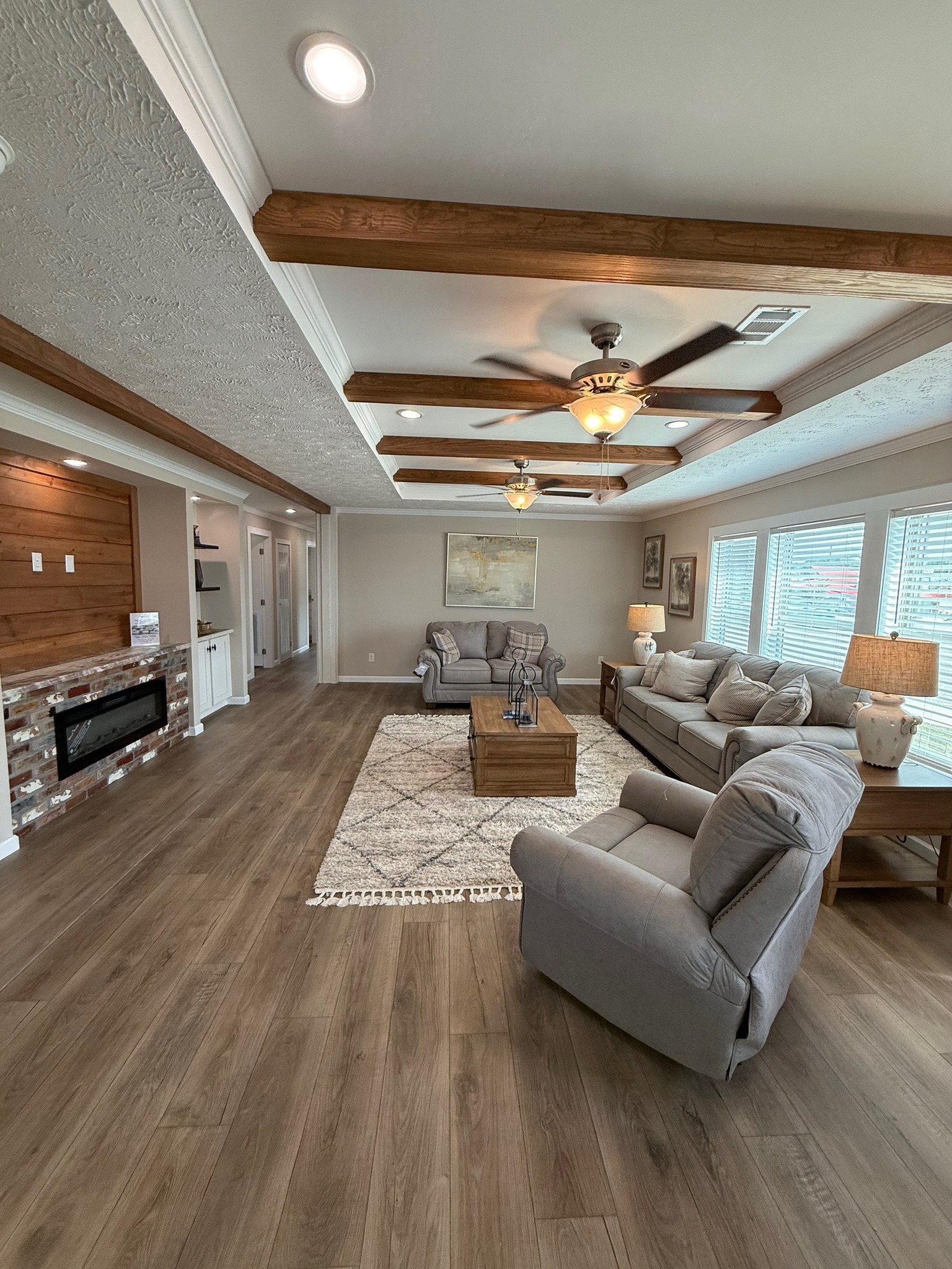 Modern living room with wood floor, gray sofas, and a cozy armchair. Exposed beams and ceiling fan add warmth. Large windows provide natural light.