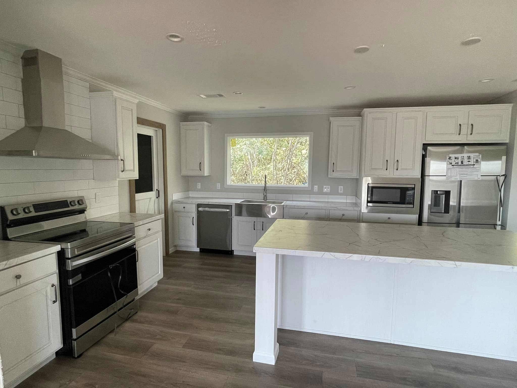 Modern kitchen with light wood flooring, stainless steel appliances, and white cabinets. A large island faces a window with a view of greenery.