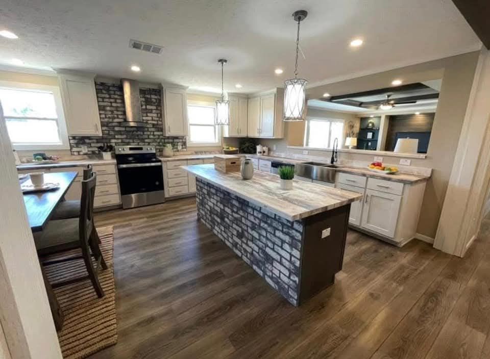 Spacious kitchen with wood floors, featuring a brick island, pendant lights, and a mix of stainless steel appliances. A dining area is visible on the left.