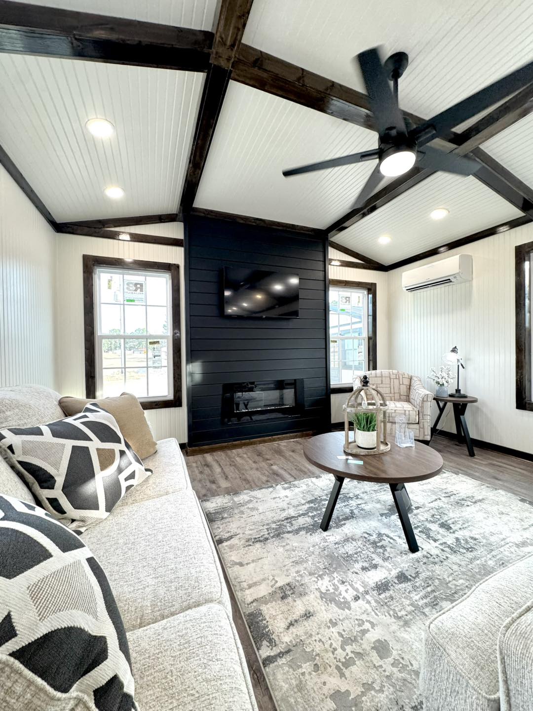 Cozy living room with vaulted ceiling, dark wooden beams, and a ceiling fan. Features a textured rug, round wooden coffee table, and modern black fireplace.