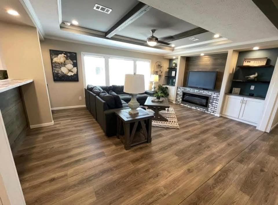 Modern living room with wood flooring, dark sectional sofa, and large TV. Ceiling fan above, natural light streams from windows on the left. Cozy and spacious ambiance.