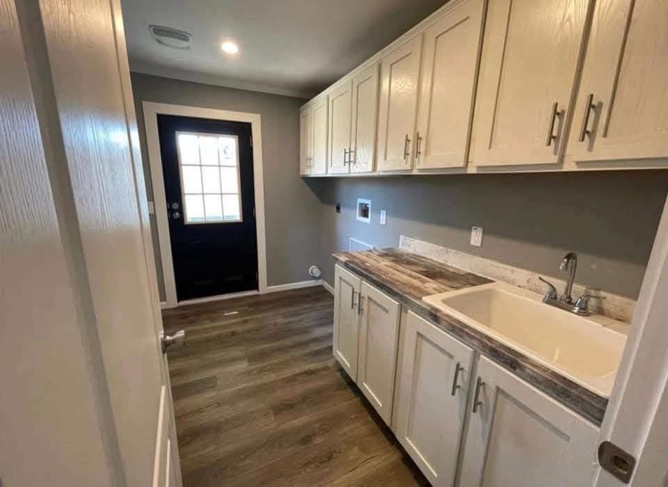 Laundry room with white cabinets and a large sink on the right, wood floor, gray walls, and a black door with a glass window. Bright and tidy ambiance.