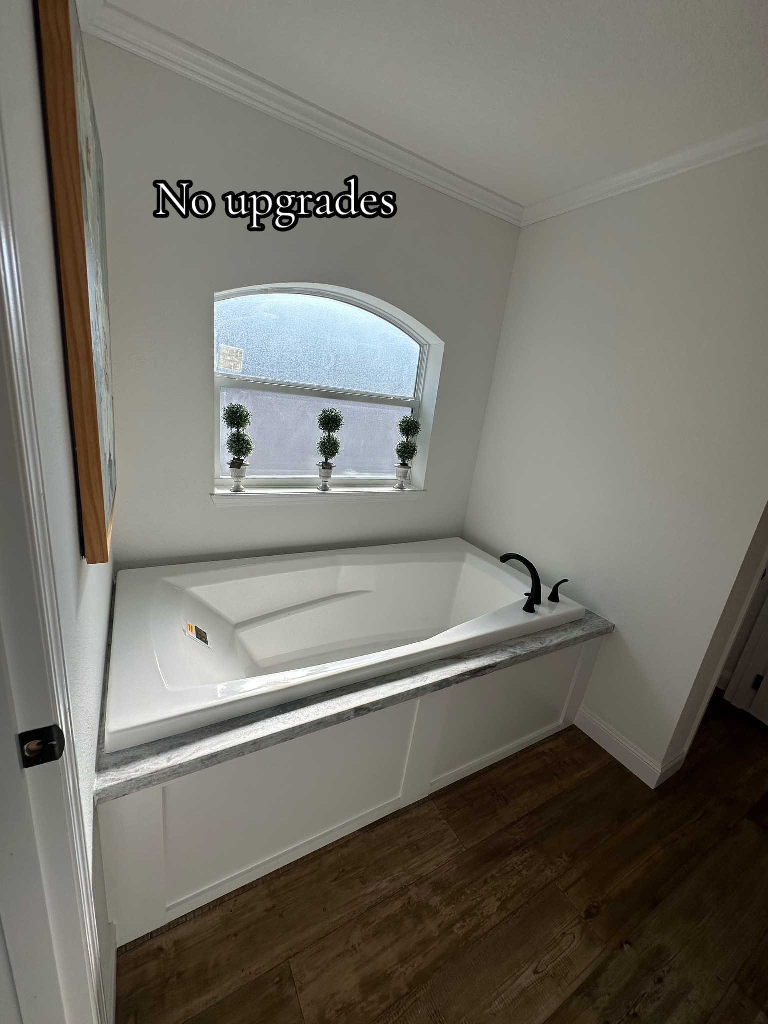 Simple bathroom with white tub under a frosted window. Window sill has small potted plants. Text reads "No upgrades." Room has wooden floors.