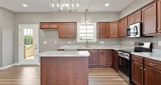 Spacious kitchen with brown wooden cabinets, center island, stainless steel appliances, and a light wooden floor. A door leads to an outdoor deck.