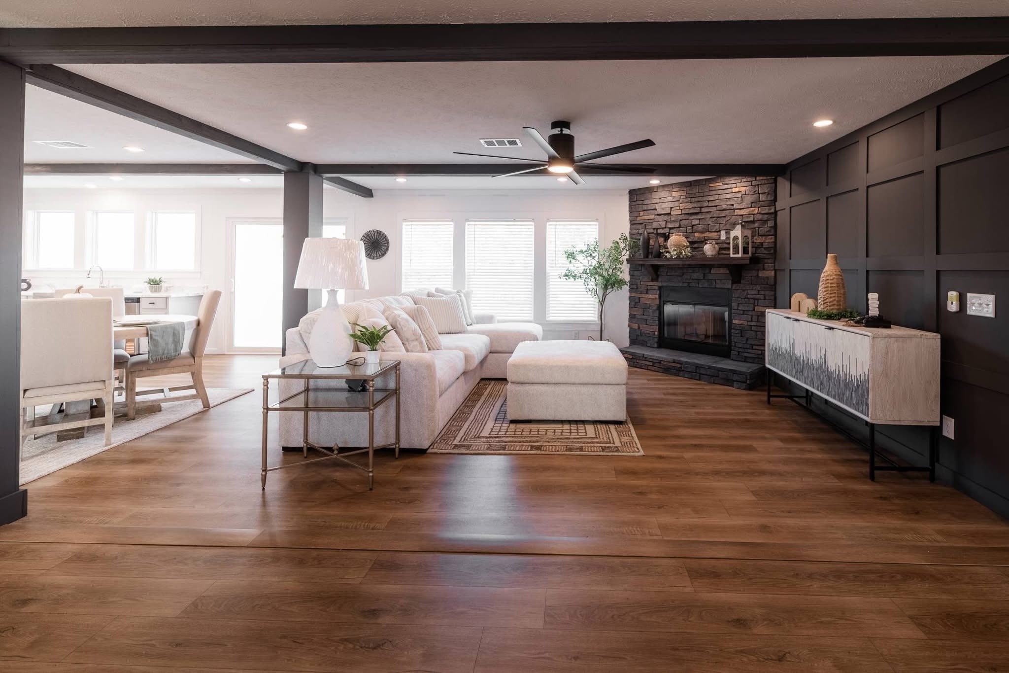 Spacious living room with light wood floors, white sectional sofa, and a stone fireplace. Large windows brighten the space, creating a cozy, modern feel.