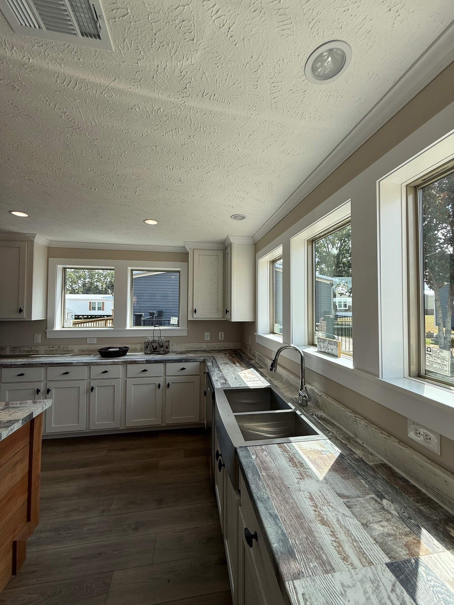 Bright kitchen with rustic, wood-patterned countertops, white cabinets, and stainless steel sink. Sunlight streams through large windows, creating a warm, inviting atmosphere.