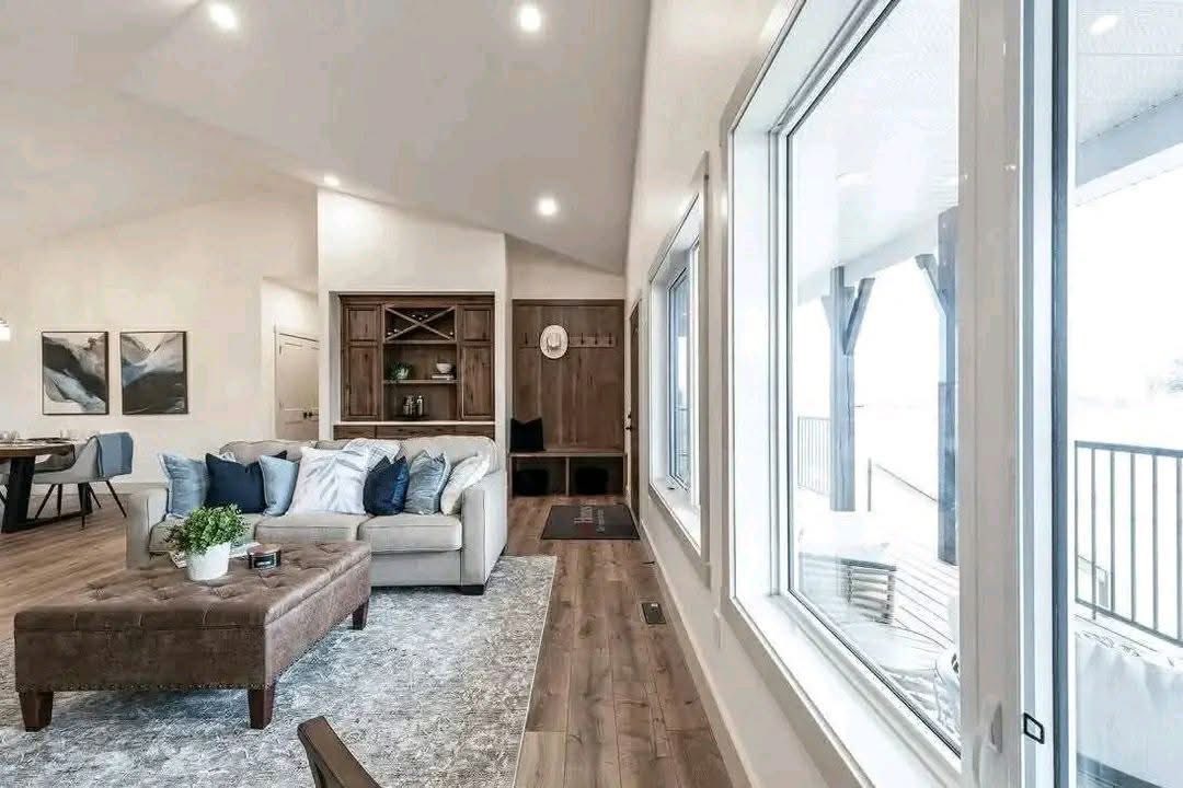 Spacious living room with large windows, beige sofa adorned with blue cushions, and a brown ottoman on a gray rug. Light wood floors and soft lighting.