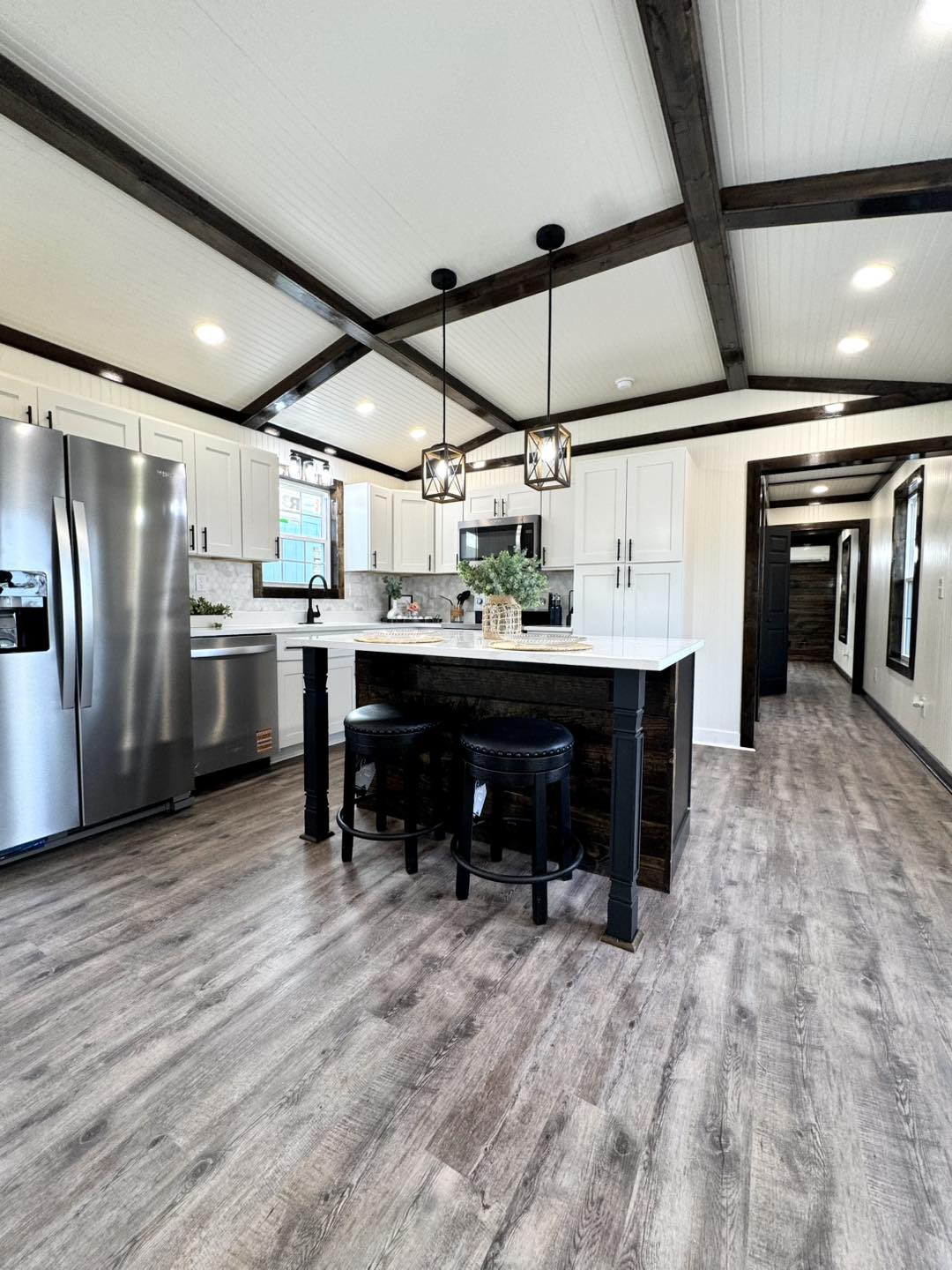 Modern kitchen with light wood flooring, white cabinets, and stainless steel appliances. An island with black stools and pendant lights adds elegance.