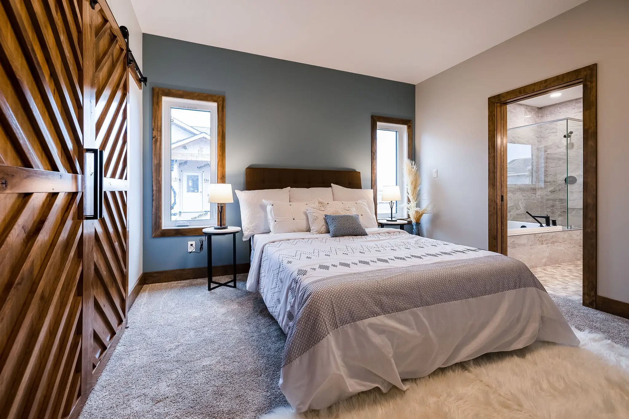 Modern bedroom with a cozy ambiance. Features a bed with white and gray bedding, wooden sliding barn door, blue accent wall, and an en-suite bathroom.