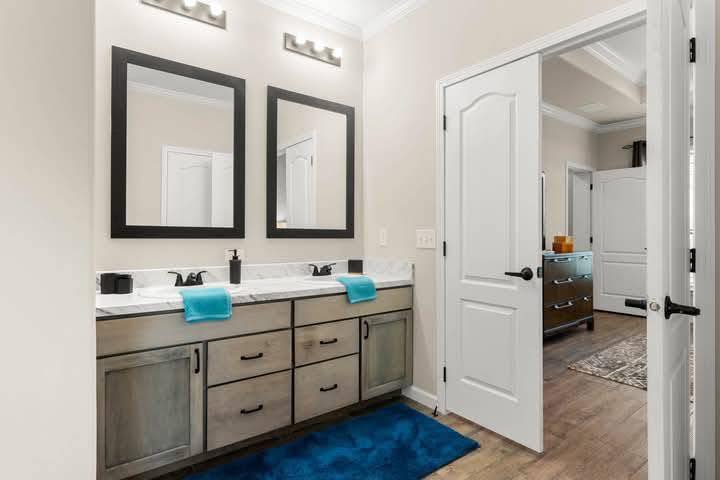 Modern bathroom with two mirrors above a double vanity, featuring wooden cabinets, black fixtures, and blue accents. Open door leads to adjoining room.