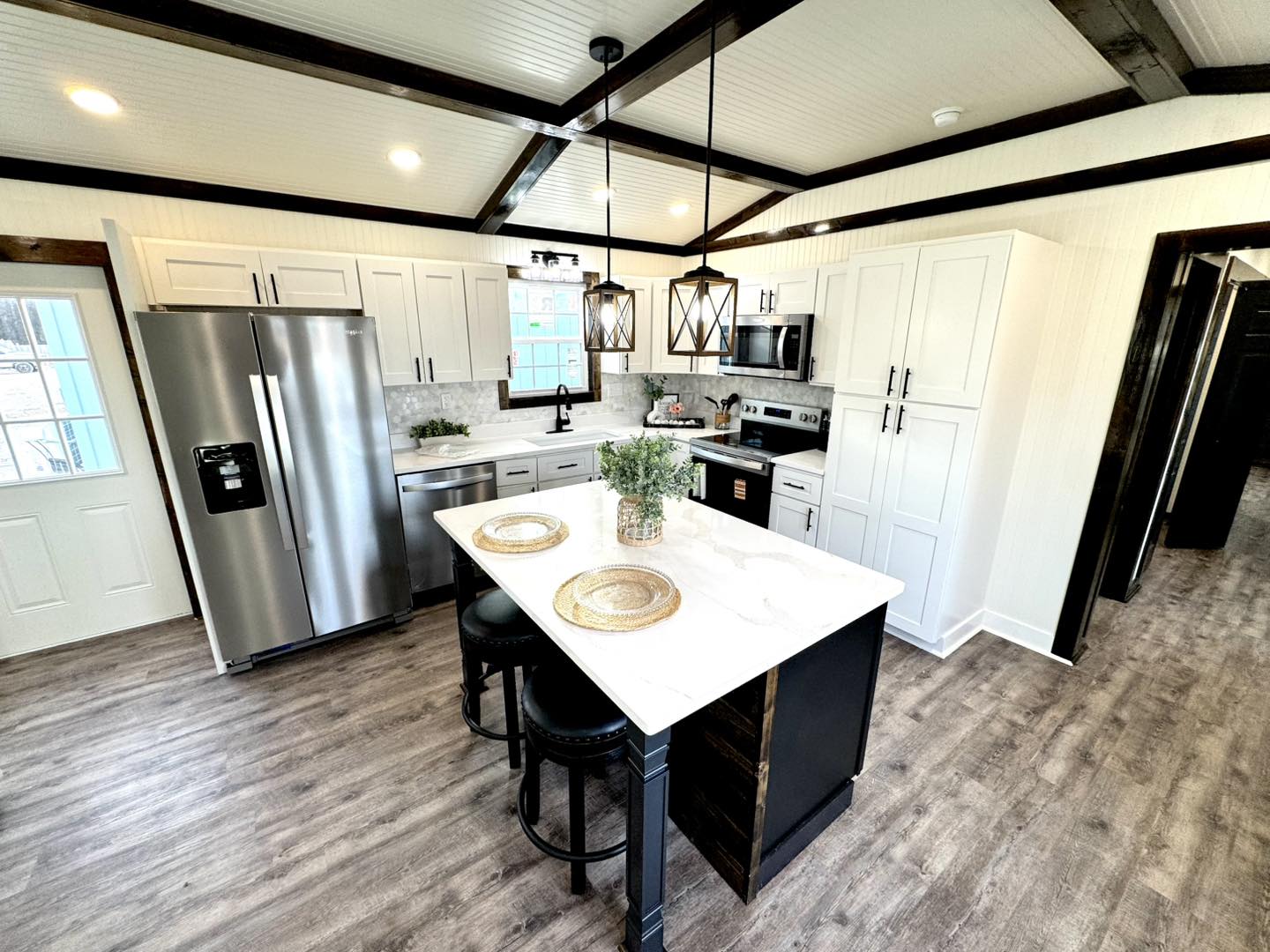 Modern kitchen with white cabinets, stainless steel appliances, and a marble island with barstools. Wooden floors and exposed beams add warmth.