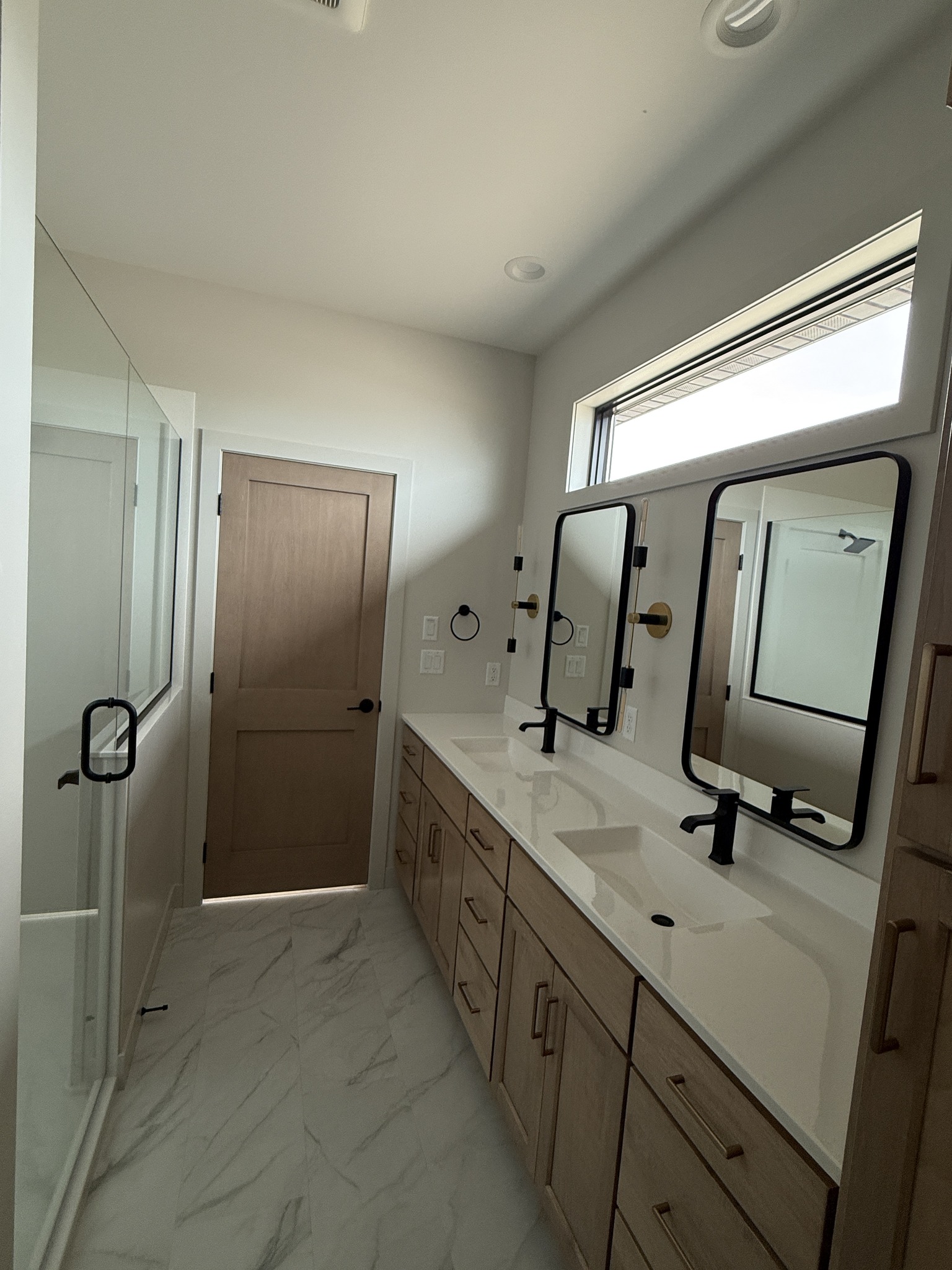 Modern bathroom with white marble floor, light wood cabinets, double sinks, and black-framed mirrors. A wooden door and glass shower add elegance.