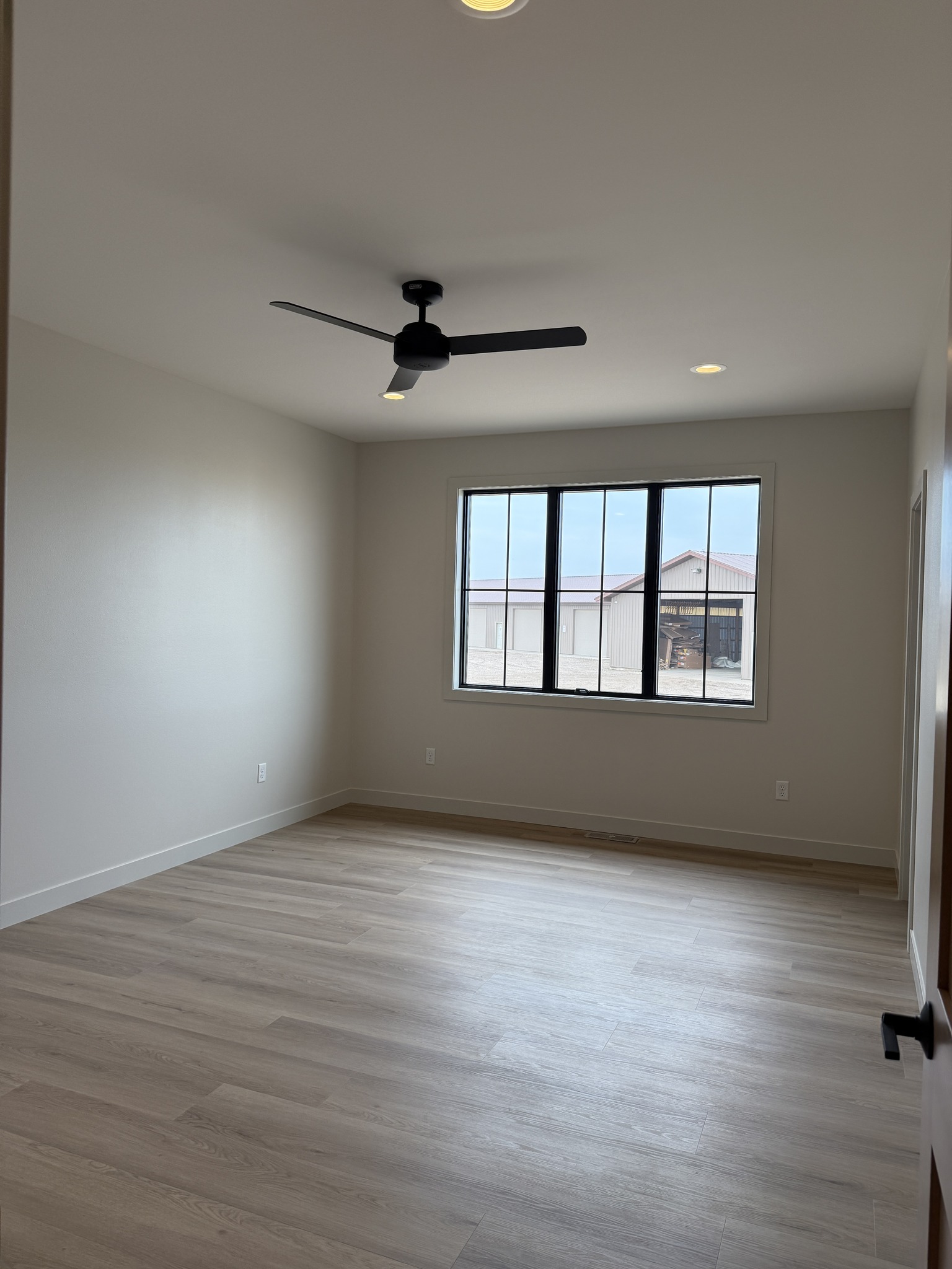 Empty room with light wood flooring, beige walls, and a large window showing an industrial building outside. Ceiling fan and recessed lighting convey modern simplicity.