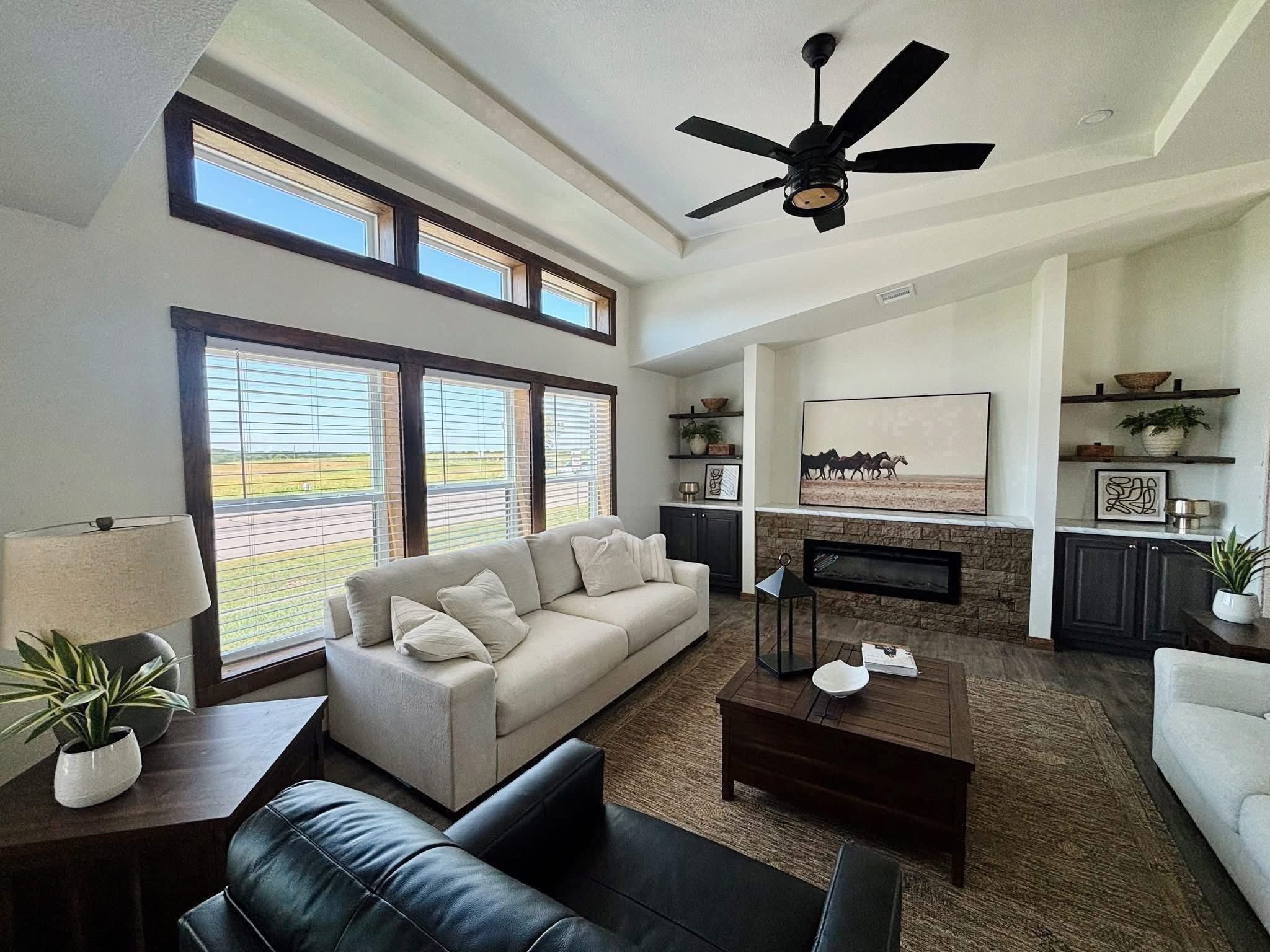 Modern living room with large windows, cream sofa, and black armchair. A ceiling fan, wall art of horses, and plants create a cozy, elegant space.