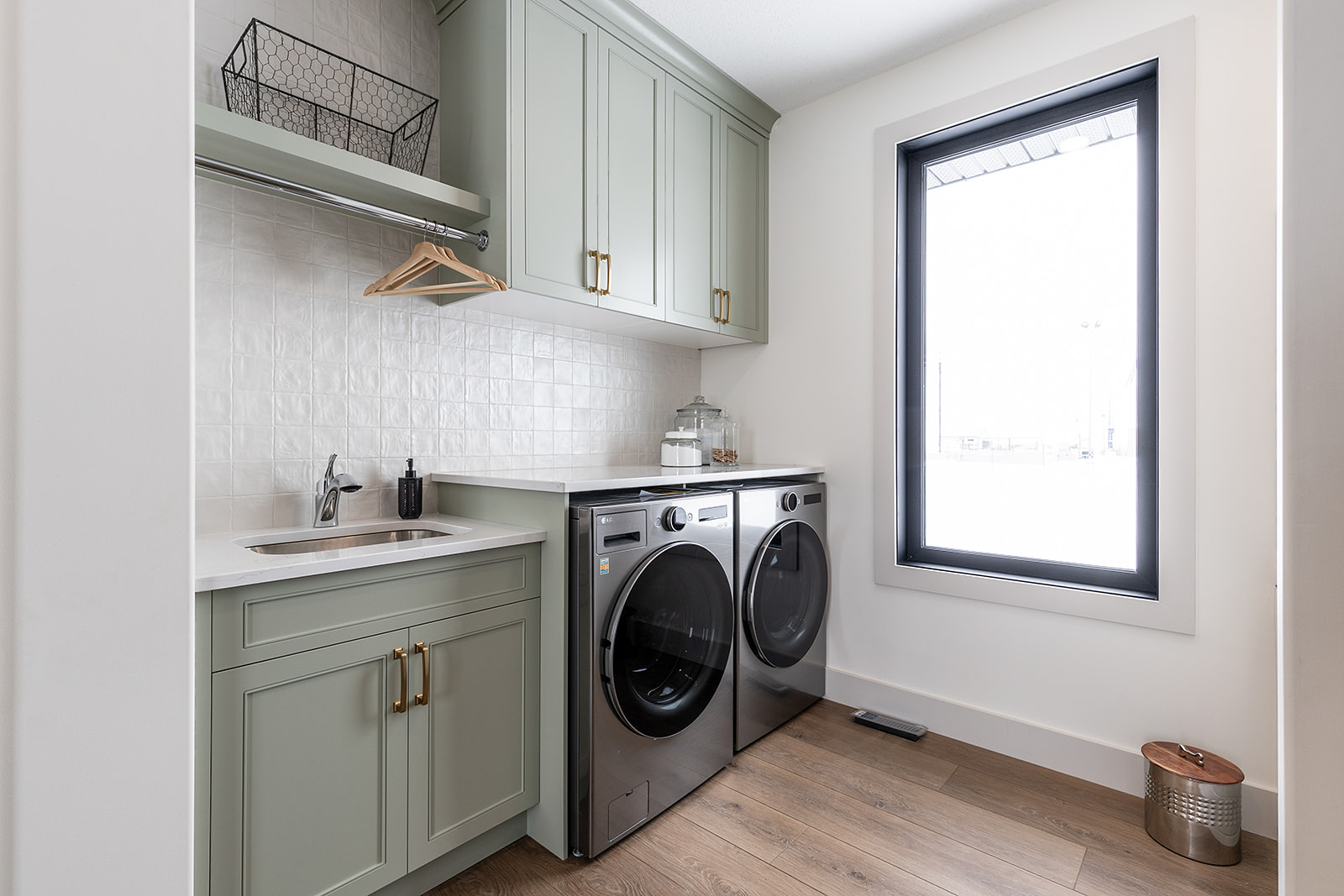 Modern laundry room with mint green cabinets, a stainless steel sink, washer and dryer, light wood flooring, and a large window for natural light.