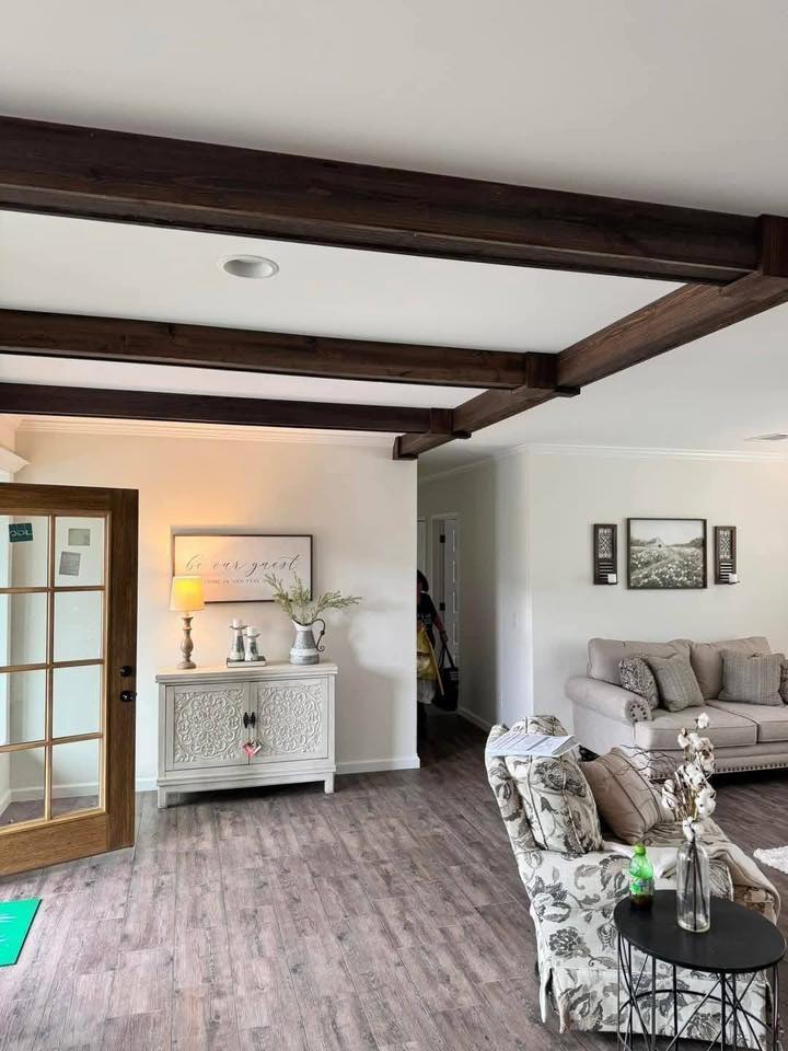 Modern living room with wooden beams, a cozy sofa, floral armchair, and a white cabinet. Soft lighting creates a warm, inviting atmosphere.