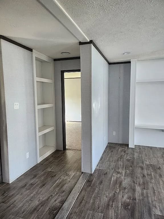 Simple interior with grayish wood flooring, light gray walls, white built-in shelves, and a doorway leading to a softly lit carpeted room. Clean, minimalist feel.