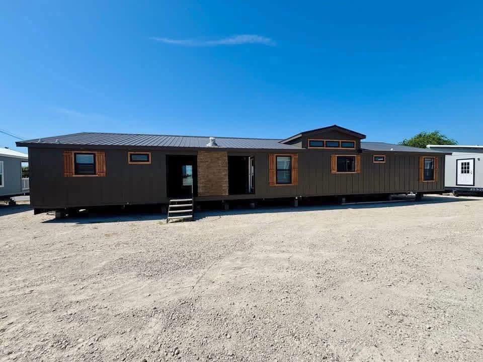 Single-story manufactured home with dark exterior, wooden accents, and large windows. It stands on a gravel lot under a clear blue sky.