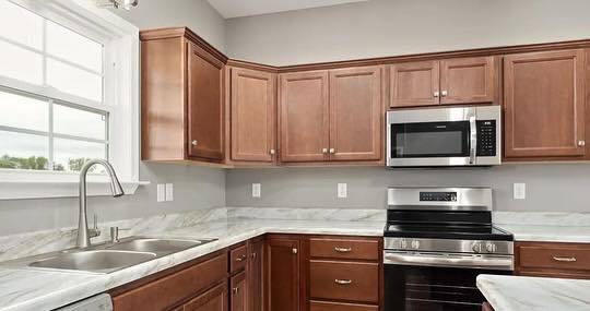 Modern kitchen with brown wooden cabinets, stainless steel microwave and oven, marble countertops, dual sink under a window, and neutral walls.