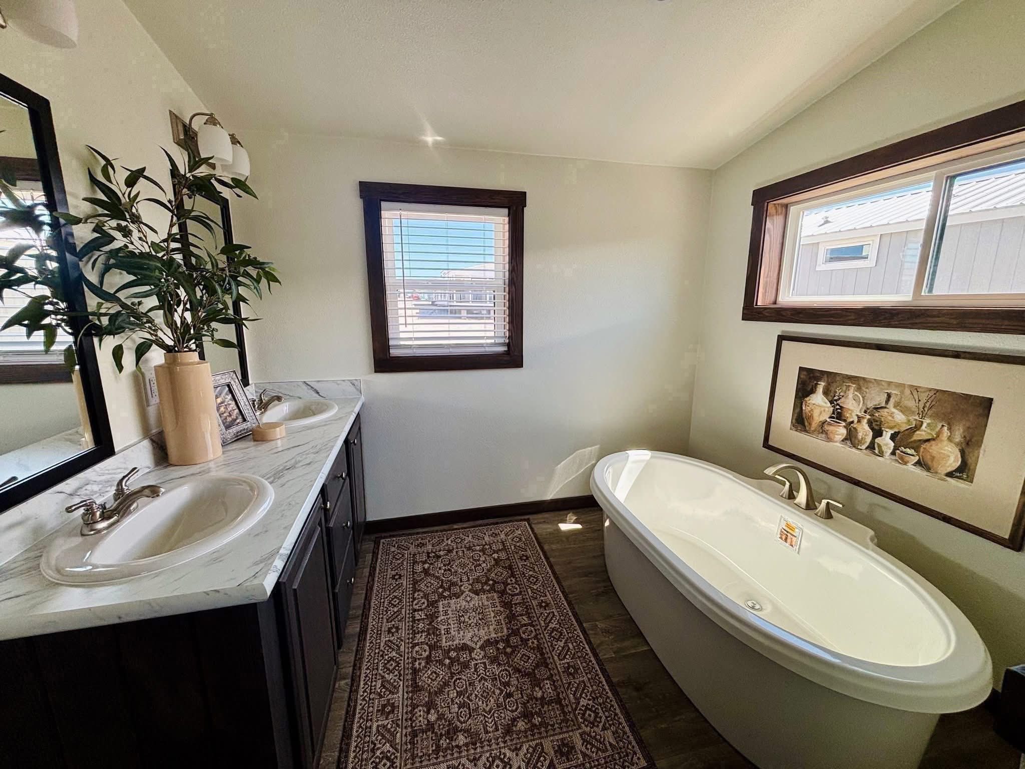 Bright bathroom with a modern design featuring a freestanding bathtub under a window, a marble-topped sink, a decorative rug, and framed art.