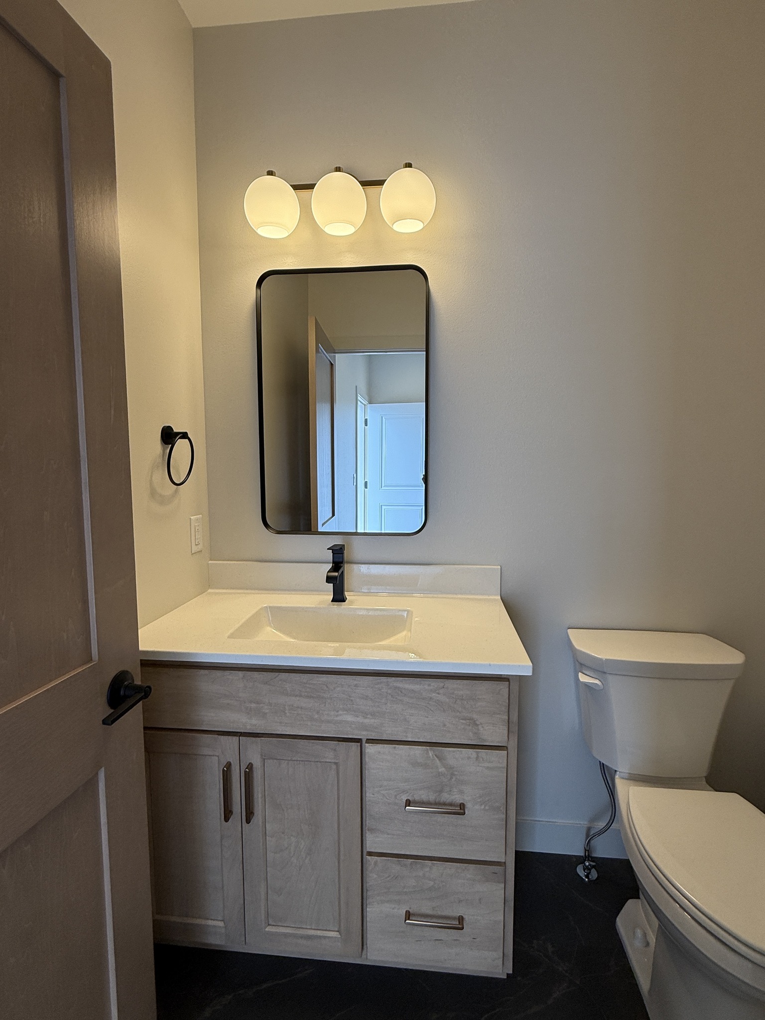 Modern bathroom with a wooden vanity, black faucet, and rectangular mirror. Three globe lights above. White toilet adjacent on dark flooring. Calm ambiance.