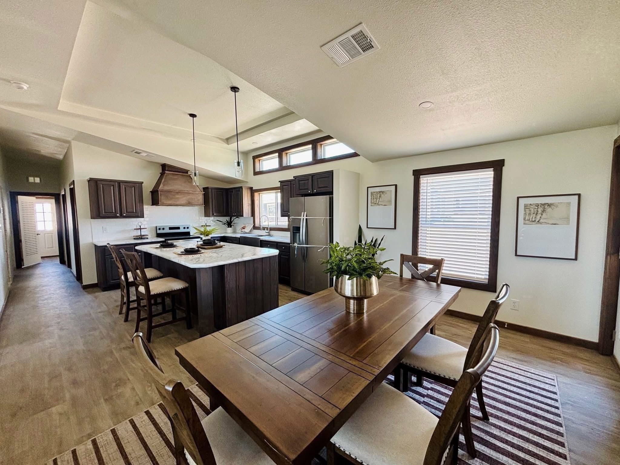 Spacious kitchen and dining area with high ceilings, dark wood cabinets, and a large table. Bright natural light streams in through a window.
