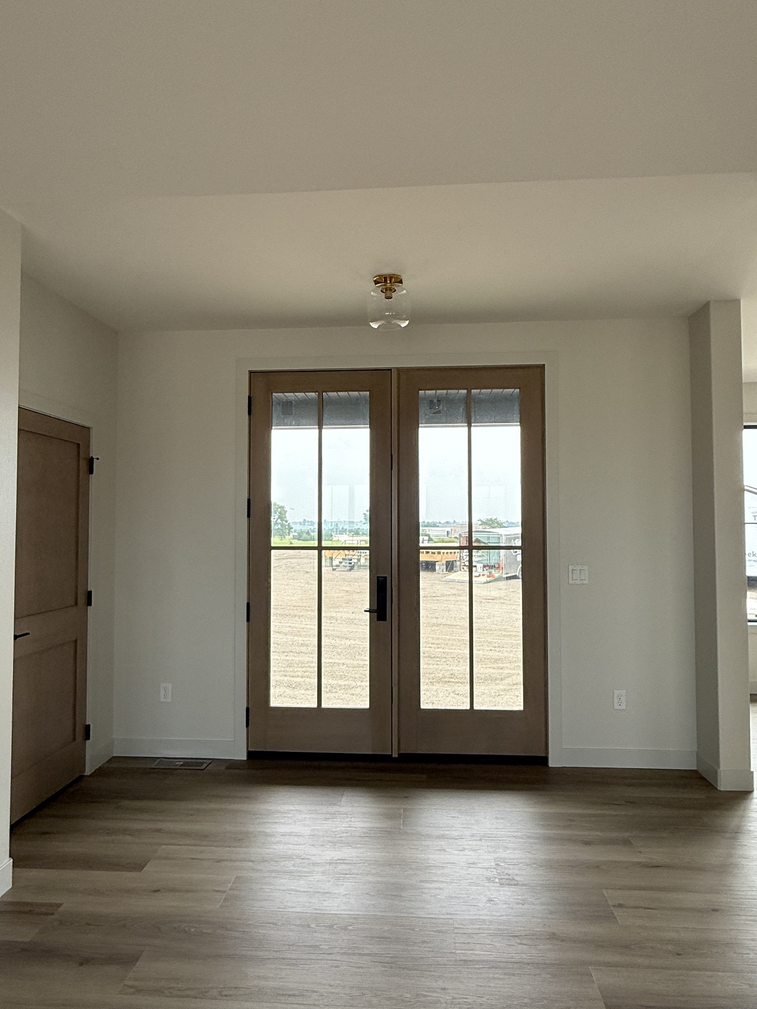 Minimalist interior with glass double doors framed in wood, leading to a construction site view. Hardwood flooring adds warmth; room feels bright and open.