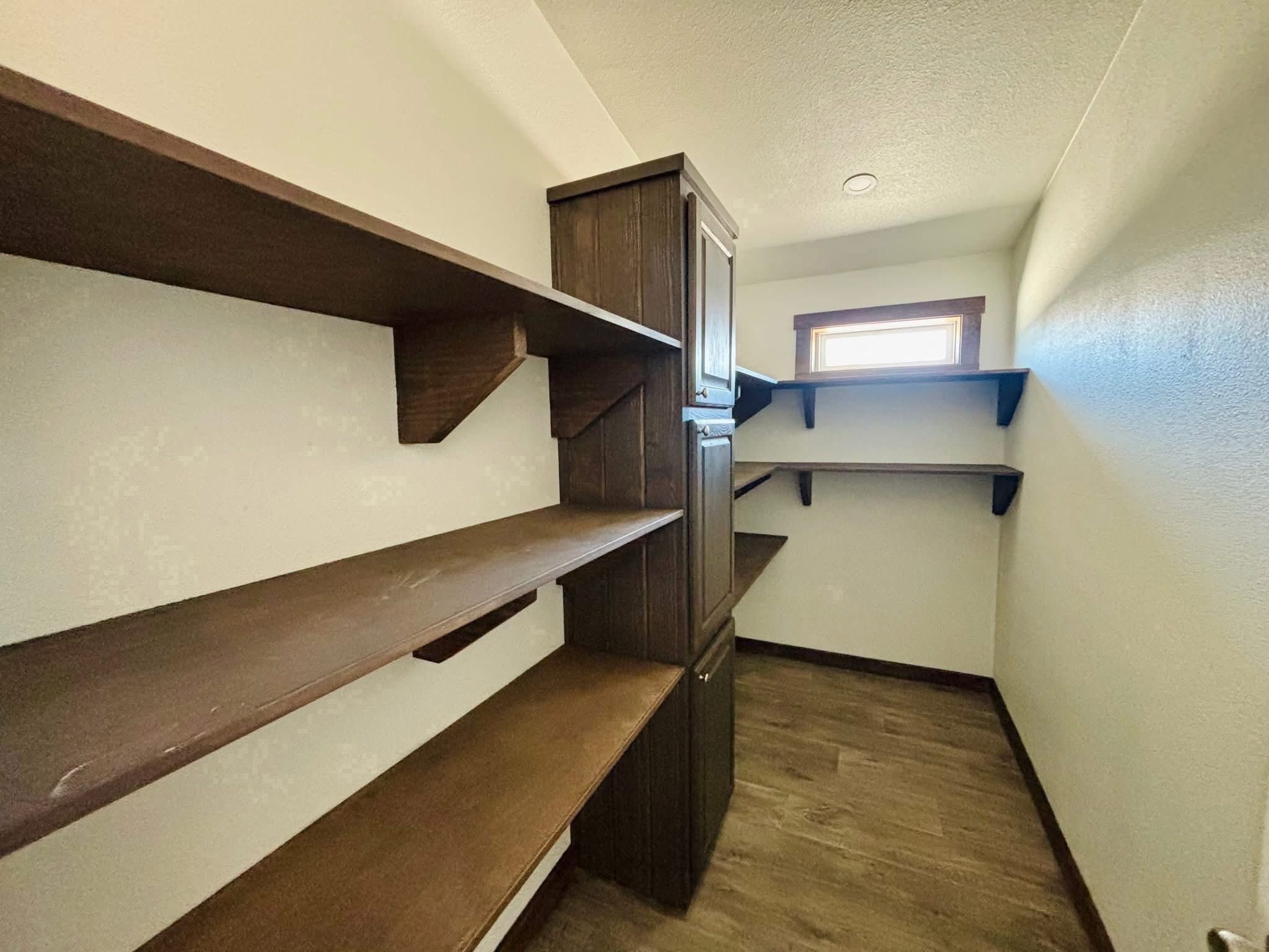 Narrow pantry with dark wooden shelves and cabinets on white walls. A small window allows natural light, creating a cozy, organized atmosphere.